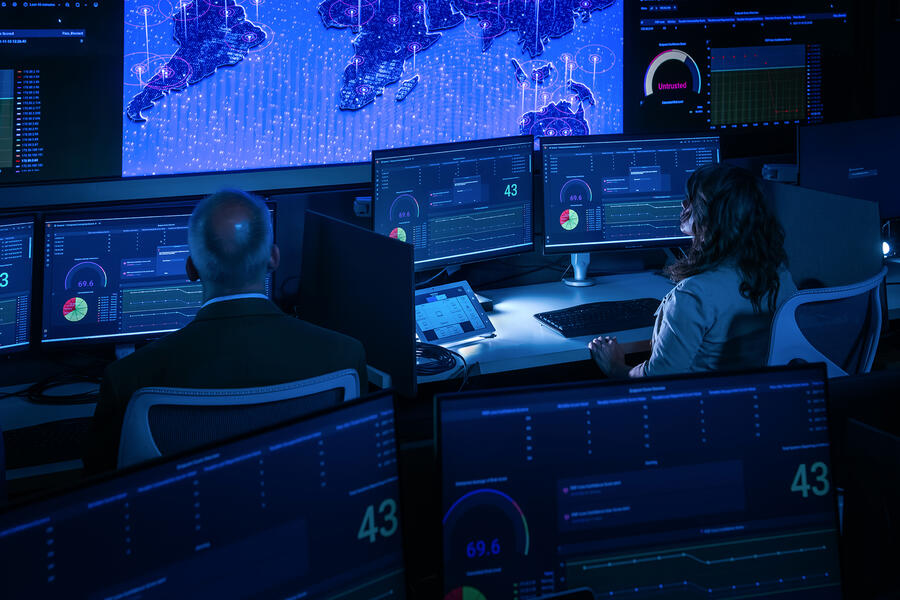 Two analysts monitor cybersecurity dashboards and global network activity on large screens in a high-tech operations center.