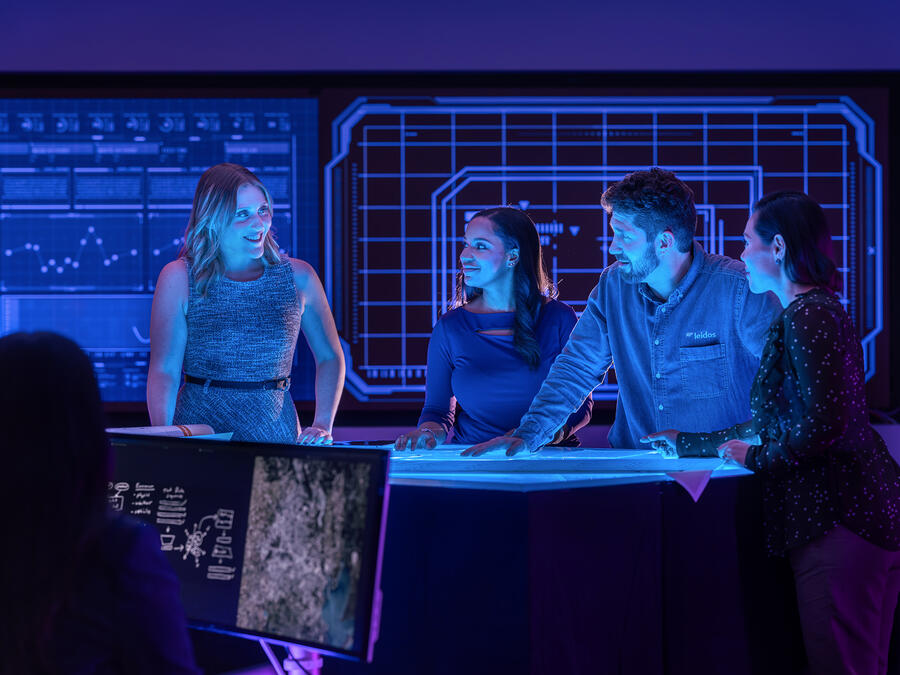 A team of professionals collaborates around a digital workspace, reviewing data and plans in a technology operations center illuminated by large interactive displays.