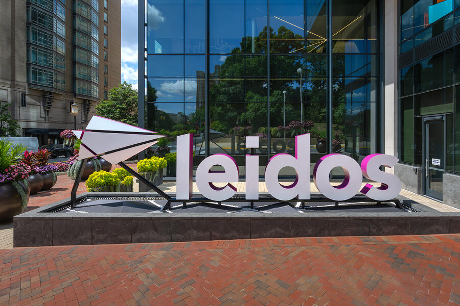A large Leidos logo sign stands outside a modern glass office building, surrounded by landscaped greenery and brick walkways.