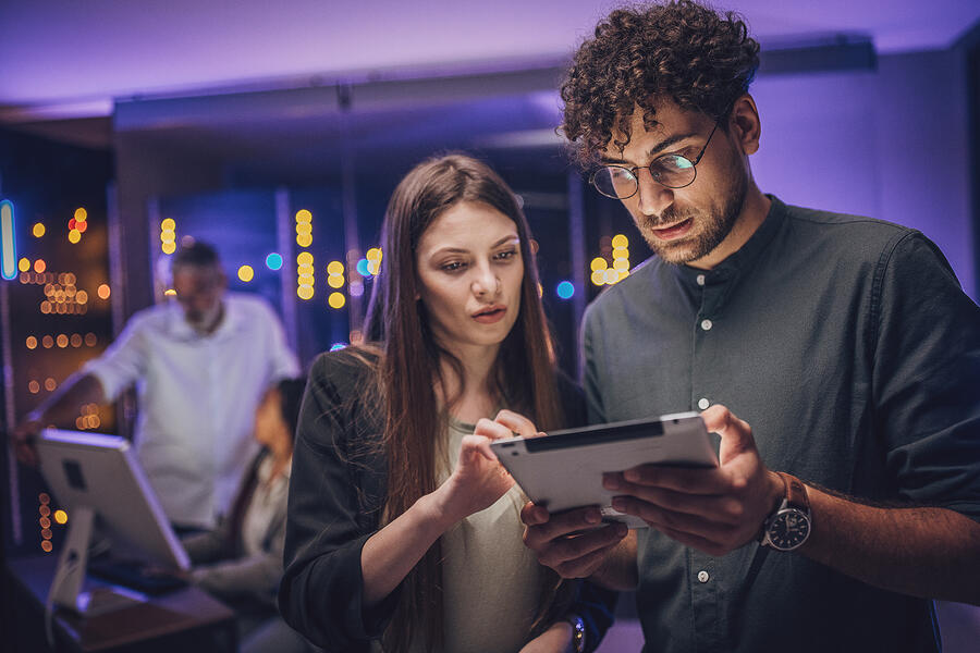 Two professionals collaborate over a tablet in a technology-focused workspace, reviewing information together under ambient purple lighting.
