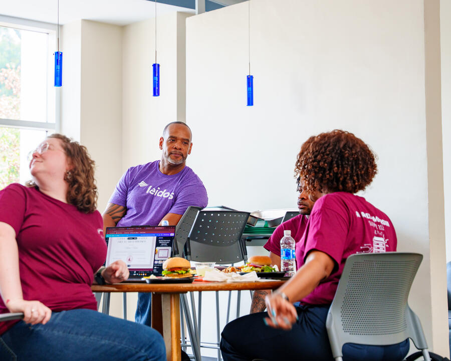 Leidos employee speaks with students during an AI summer immersion program as participants gather around a table for discussion and lunch.