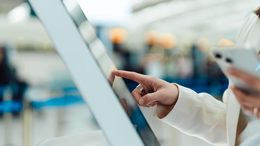 Close-up of a traveler using a touchscreen self-service kiosk while holding a smartphone, illustrating digital identity verification and contactless passenger processing at an airport.