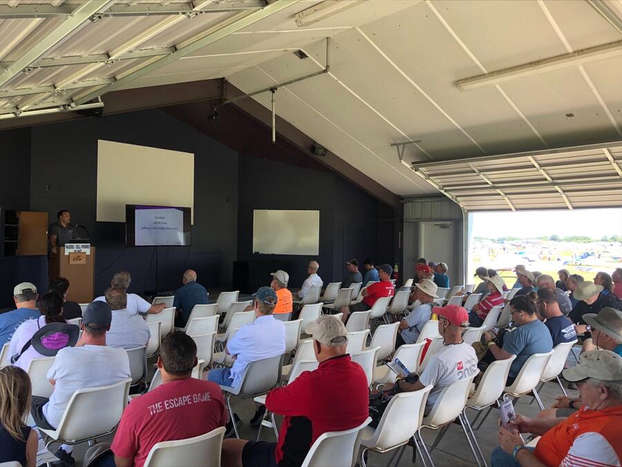 Attendees sit in rows of chairs inside an open-air hangar while a presenter delivers a talk using a podium and presentation screen during an aviation or technology event.