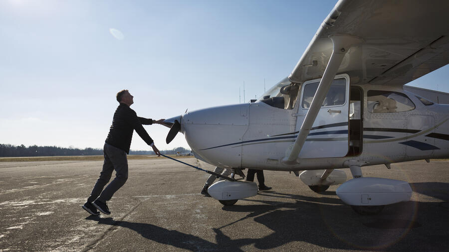 A person pushes a small single-engine aircraft across an airport ramp before flight, with another individual boarding through the open cabin door under a clear sky.