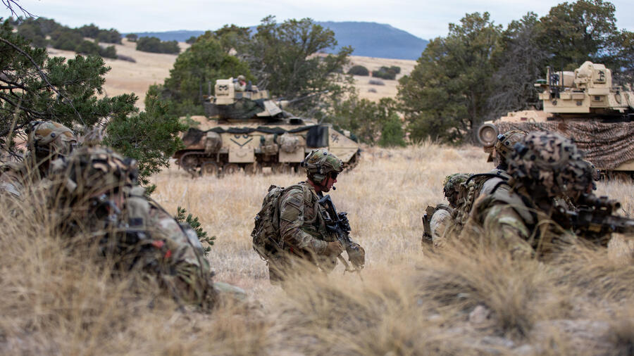 Soldiers move through rugged terrain during a military training exercise, with armored combat vehicles providing support in the background.
