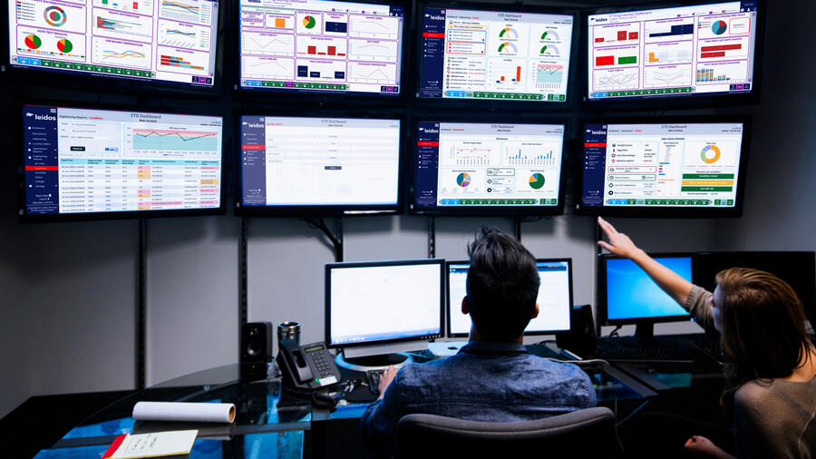 Two professionals monitor operational dashboards on multiple large displays, reviewing performance metrics, analytics, and system status in a technology operations center.