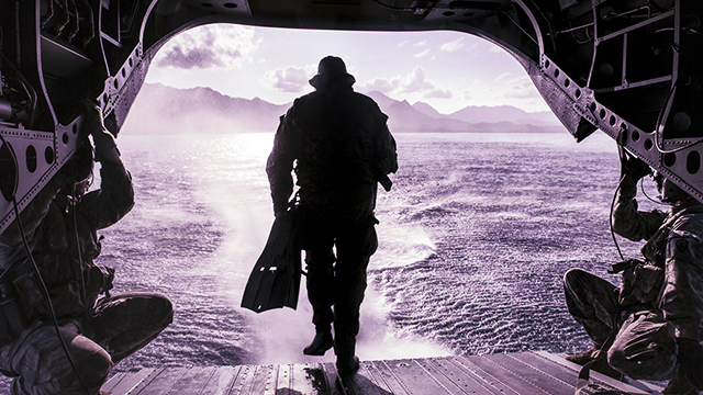 A service member exits the rear ramp of a military aircraft over open water during a maritime operation, with mountains visible in the distance and other personnel seated inside the aircraft.