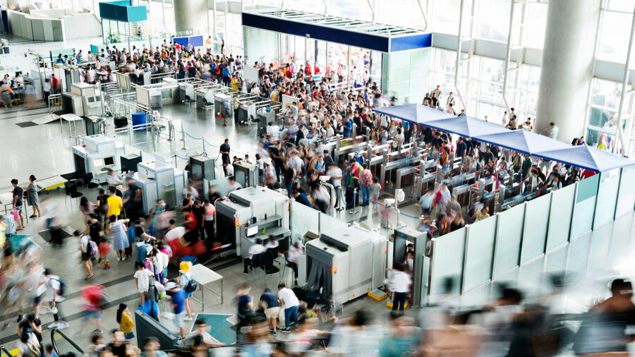 Crowds of travelers move through security screening checkpoints and queues inside a busy airport terminal.