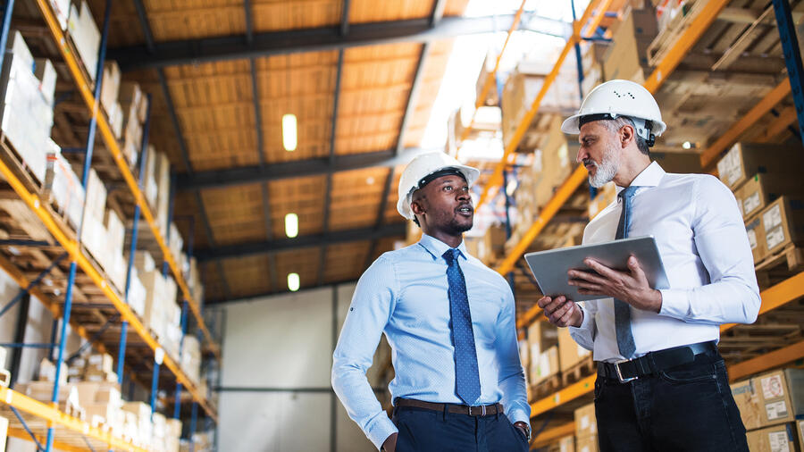 Two professionals wearing hard hats discuss operations while reviewing information on a tablet in a warehouse.