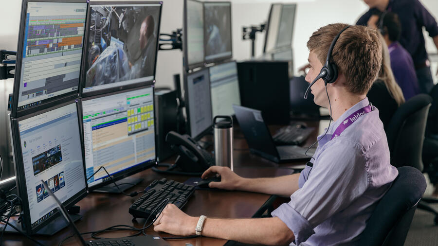 Leidos employee wearing a headset monitors multiple computer screens displaying operational data and video feeds.