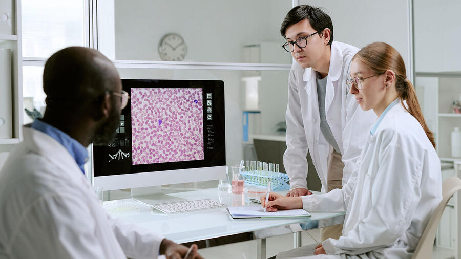 Three laboratory researchers analyze a magnified image of blood cells displayed on a computer monitor.