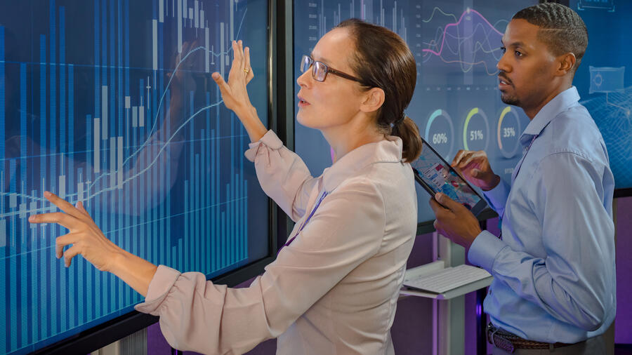 Two professionals analyze data visualizations on large digital displays, with one pointing to a chart while the other holds a tablet.