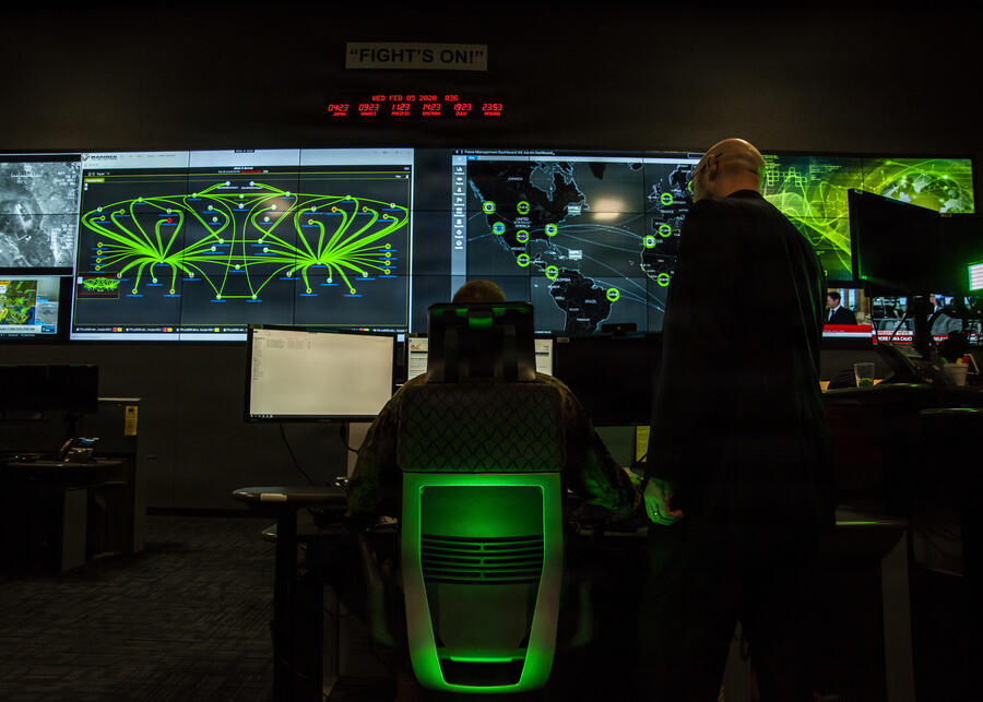 Two people facing away looking at large computer and TV screens with various data feeds in a dark room
