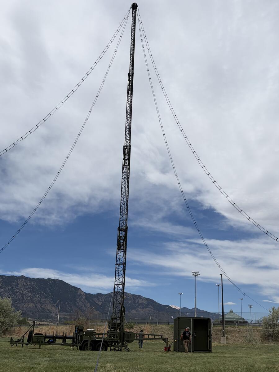 A lattice-style metal tower supported by metal feet at the base on the ground with four wire lines coming from the top to the ground - this is ALPS (Army Long-Range Persistent Surveillance) system is a passive, long-range aerial surveillance sensor developed by Leidos