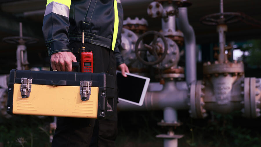Industrial worker in high-visibility clothing carries a toolbox and tablet beside large pipes and valves at an industrial facility.