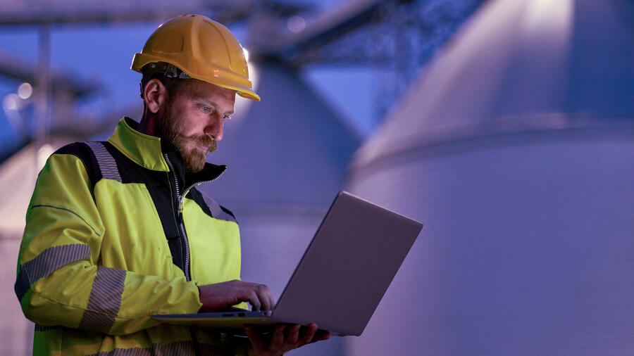 Engineer wearing a yellow hard hat and high-visibility jacket uses a laptop inside an industrial facility.