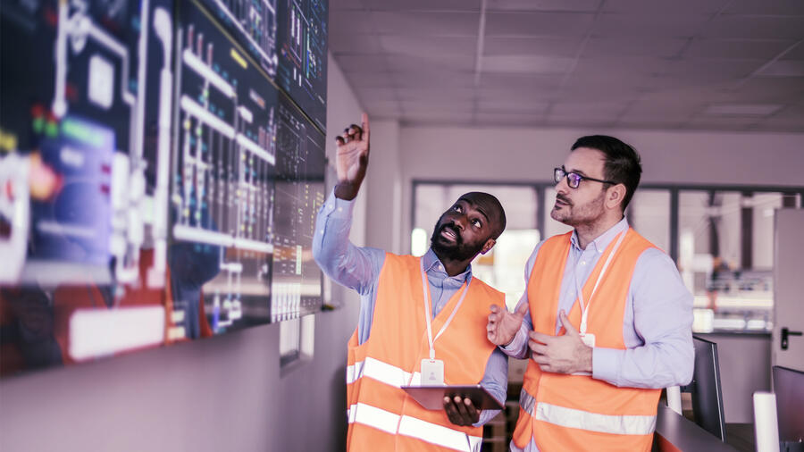 Two engineers wearing orange high-visibility vests review operational data on large digital displays, with one pointing at the screen while holding a tablet.