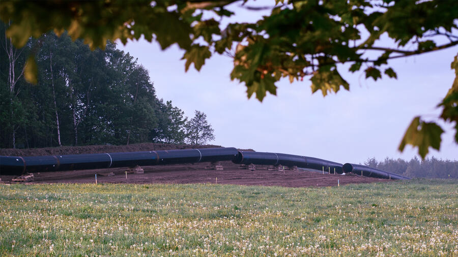 Large black gas pipeline sections stretch across a grassy field bordered by trees, with wildflowers in the foreground.