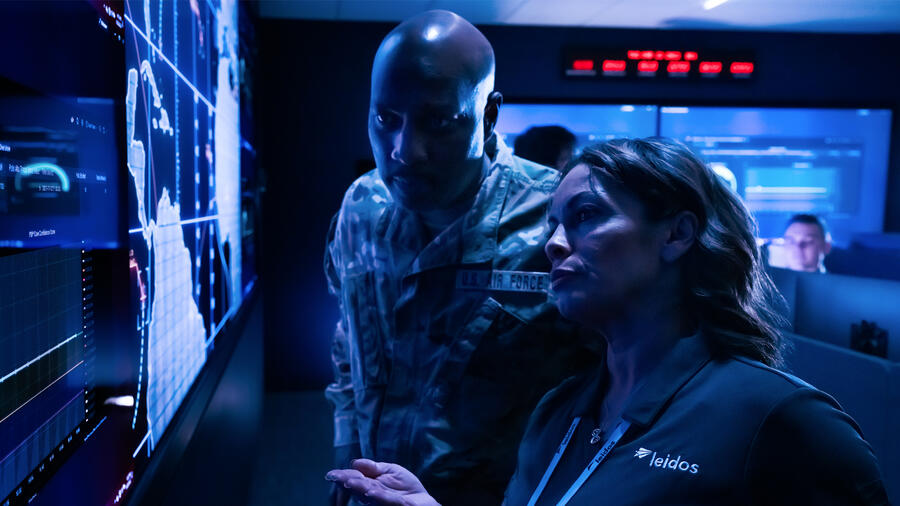U.S. Air Force service member and Leidos employee analyze data on large digital displays in a military cyber operations center.