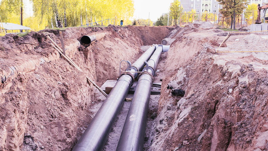 Two large metal pipelines run through an excavated trench at a construction site, surrounded by earth and construction materials.
