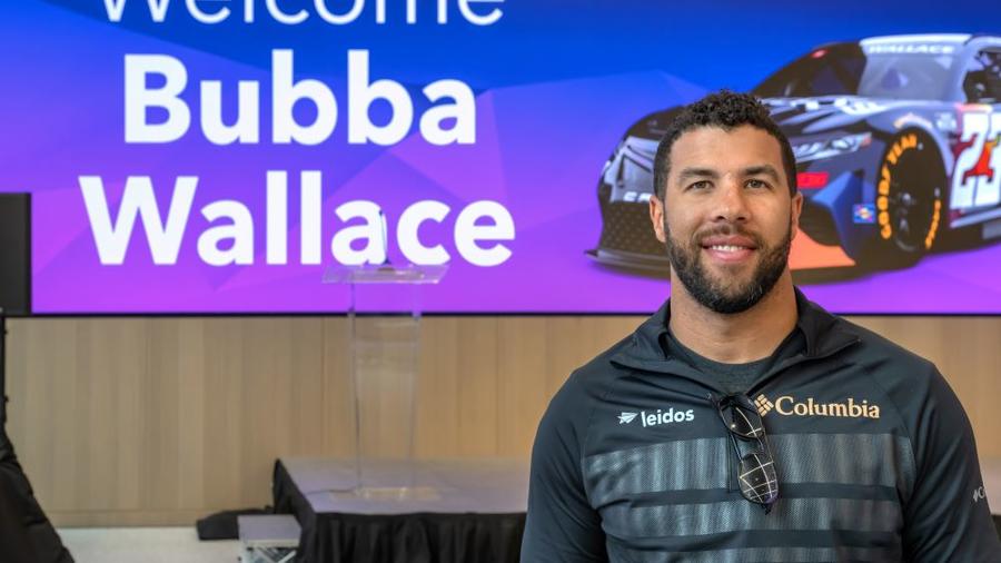 Bubba Wallace posing in front of a picture of his race car at Leidos Global Headquarters in Reston, Virginia