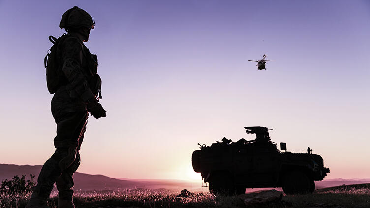Military member standing outside, alongside a vehicle and helicopter flying overhead