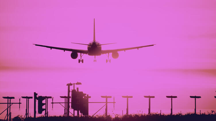 Airplane descends for landing against a vivid pink sky, silhouetted above runway lights and approach equipment.