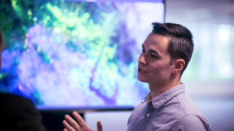 A man stands talking in front of a screen