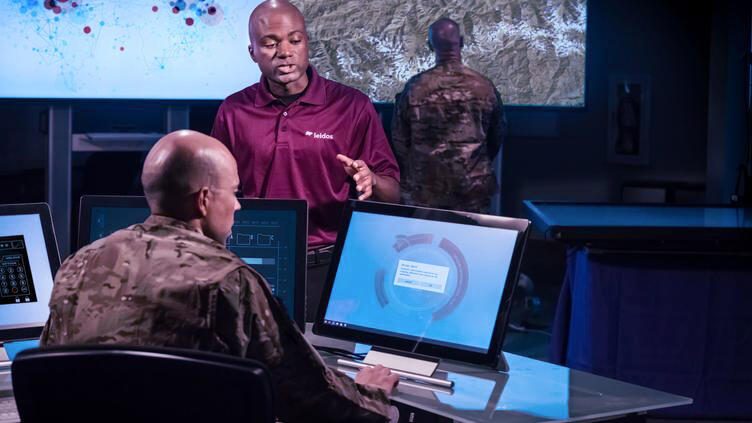 A man in a Leidos shirt talks to a military man sitting down in front of a computer screen.