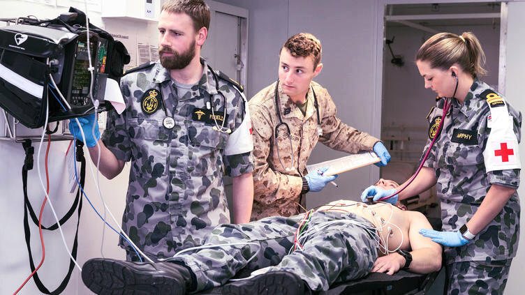One doctor examines a patient on the bed and two men look at a screen. All are in military uniforms