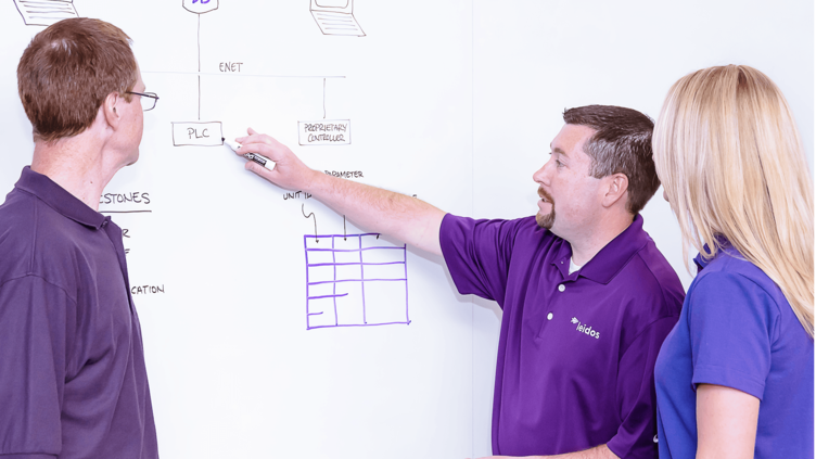 A man points to a diagram on a white board while his colleagues watch attentively